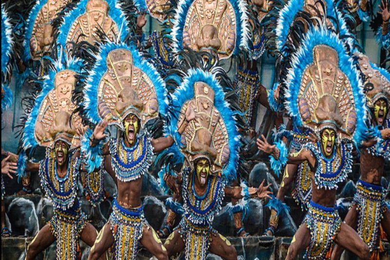 A vibrant photograph of performers in the Dinagyang Festival in the Philippines. Dancers, with dark body paint and elaborate, colorful tribal costumes and headdresses, are moving energetically in the street to the rhythm of drums. This is an example of exciting Philippine travel.