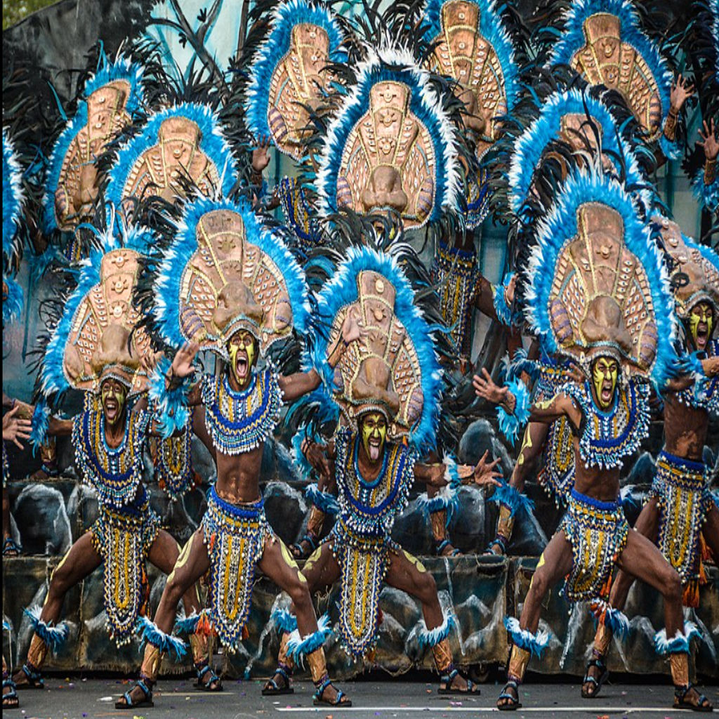 A vibrant photograph of performers in the Dinagyang Festival in the Philippines. Dancers, with dark body paint and elaborate, colorful tribal costumes and headdresses, are moving energetically in the street to the rhythm of drums. This is an example of exciting Philippine travel.